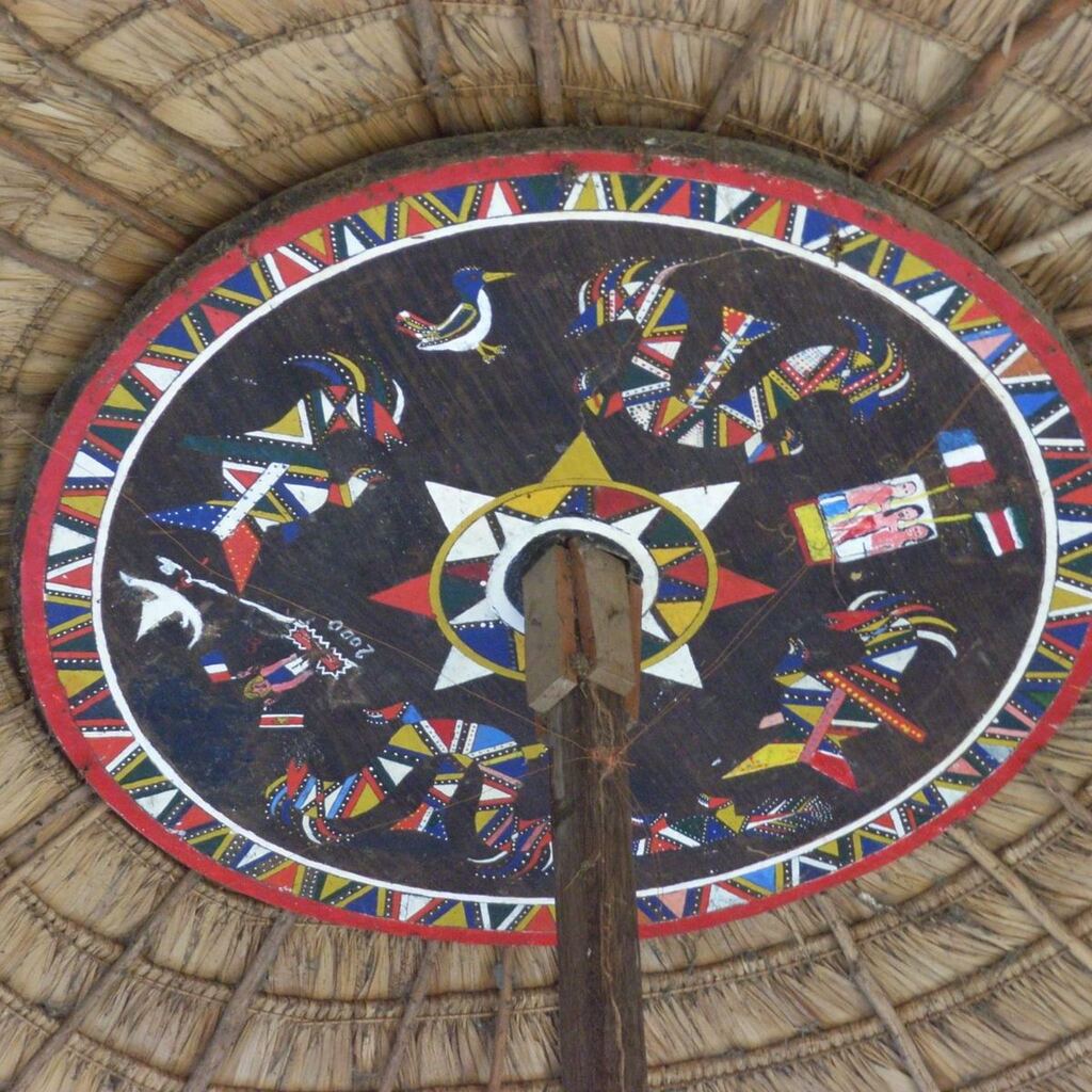 Van binnenuit gezien rieten dakconstructie met een beschilderd rond schild in tembe-motieven.